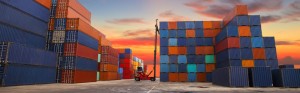 A forklift retrieves a cargo container from a tall stack of containers on a dock as the sun rises