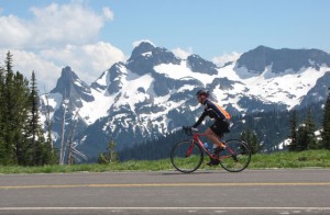 A bicyclist coasts down a mountain roadway with Mount Rainier behind him